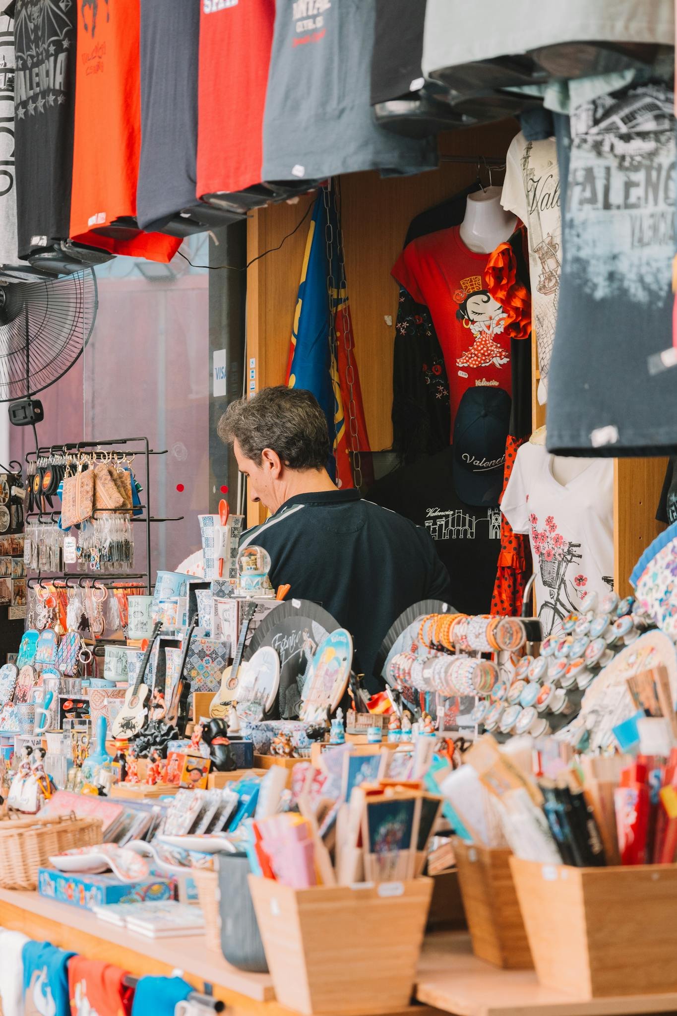 Busy market stall selling colorful souvenirs and T-shirts in an urban street setting.