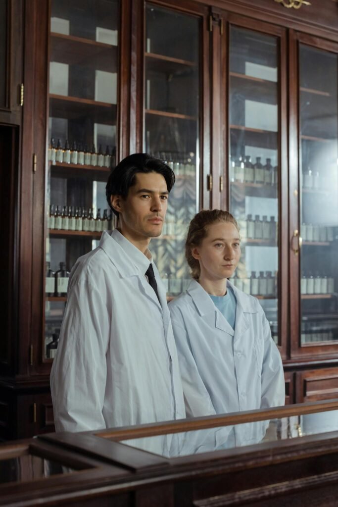 Two pharmacy employees in white coats standing by a wooden cabinet with shelves of bottles.